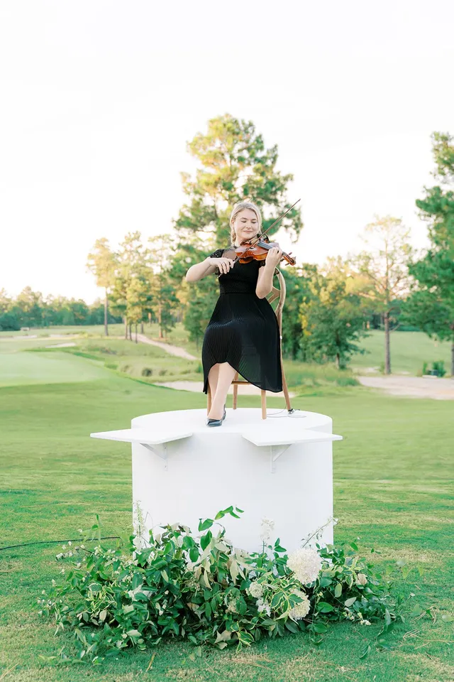 Violinist on floral-garlanded white platform at golf club in backlit golden hour light