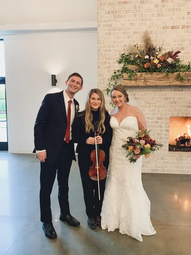 Bride, groom and violinist beside a fireplace with autumn floral mantle arrangement