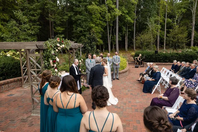 Wide ceremony panorama at brick garden venue with floral arch and seated wedding guests
