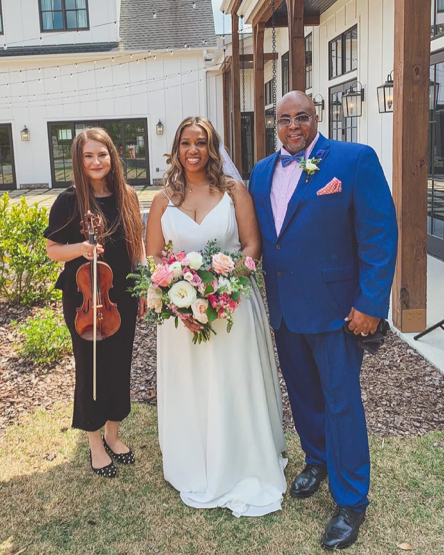 Violinist with couple at white barn with string lights and colorful floral bouquet