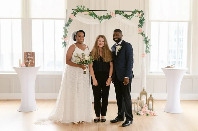 Violinist with couple in light-filled white ballroom with blush floral arch